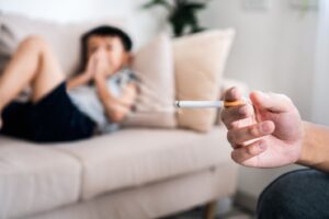 Hand holding cigarette in foreground with child lying on sofa in background.